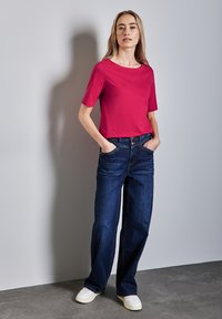 Fuchsie tričkem s krátkým rukávem v kombinaci s tmavě modrými širokými džíny, které mají uvolněný střih a klasický design s pěti kapsami, doplněné bílými teniskami.