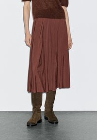 Gonna plissettata bordeaux in tessuto leggero, abbinata a stivali al ginocchio in suede marrone. L'outfit presenta una vestibilità rilassata e una drappeggiatura elegante.