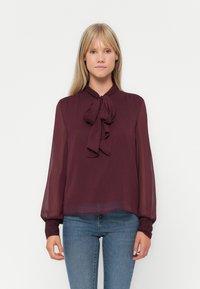 Blusa bordeaux realizzata in tessuto leggero e testurizzato, con un fiocco al collo e maniche lunghe e voluminosi. Abbinata a jeans blu.