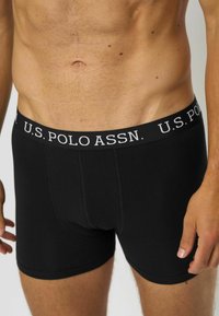 Čierne boxerky s širokým pásom, na ktorom je biely nápis "U.S. POLO ASSN.", hladká tkanina, priliehavý strih a žiadne viditeľné vzory.