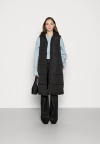 Veste sans manches noire rembourrée avec capuche, dotée d'une fermeture éclair à l'avant, portée sur un pull bleu clair, associée à un pantalon noir et un sac.