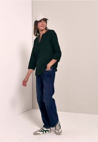 Blusa verde scuro con scollo a V e maniche a tre quarti, abbinata a jeans blue e sneaker grigi con accenti verdi. Cappellino bianco.