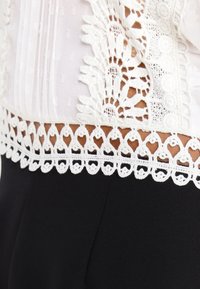 Bordure en dentelle présentant des motifs floraux complexes en blanc, fixée à un vêtement léger à la texture délicate, contrastant avec une base sombre.