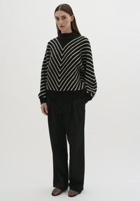 Maglione a righe bianche e nere con collo alto, caratterizzato da un motivo a zig-zag. Abbinato a pantaloni neri larghi e scarpe con tacco in rete.