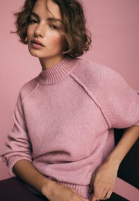 Helles pinkes Strickpullover mit einem gerippten Stehkragen und kurzen Ärmeln, mit einer weichen Textur und lockerem Schnitt. Vor einem pinken Hintergrund.