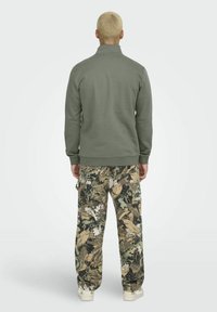 Sweatshirt verde măsliniu cu guler înalt, purtat cu pantaloni de camuflaj care au modele de frunze în nuanțe de verde, maro și negru.
