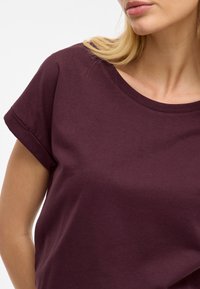 Burgundska bombažna majica s kratkimi zavihanimi rokavi in širokim izrezom. Gladka tekstura brez vzorcev ali vidnih akcentov.