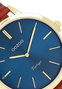 Montre ronde avec un cadran bleu, marqueurs d'heure en or et aiguilles. Bracelet en cuir marron, boîtier doré et logo "Oozoo Vintage" sur le cadran.