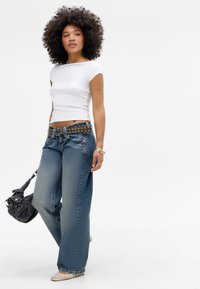 Hvid kortærmet top parret med lyseblå denim wide-leg jeans, der har lodrette striber og et spænde med nitter. Holder en mørk taske.