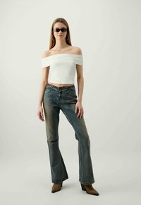 Top branco crop off-the-shoulder com textura plissada, combinado com jeans flare azul desbotado e botas de cano baixo com bico fino castanhas.
