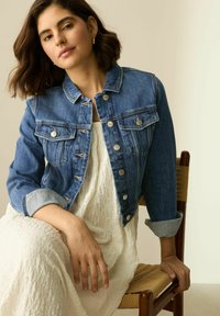 Giacca in denim di blu medio con taglio classico, tasche sul petto e dettagli in argento. Indossata su un vestito bianco a trama.