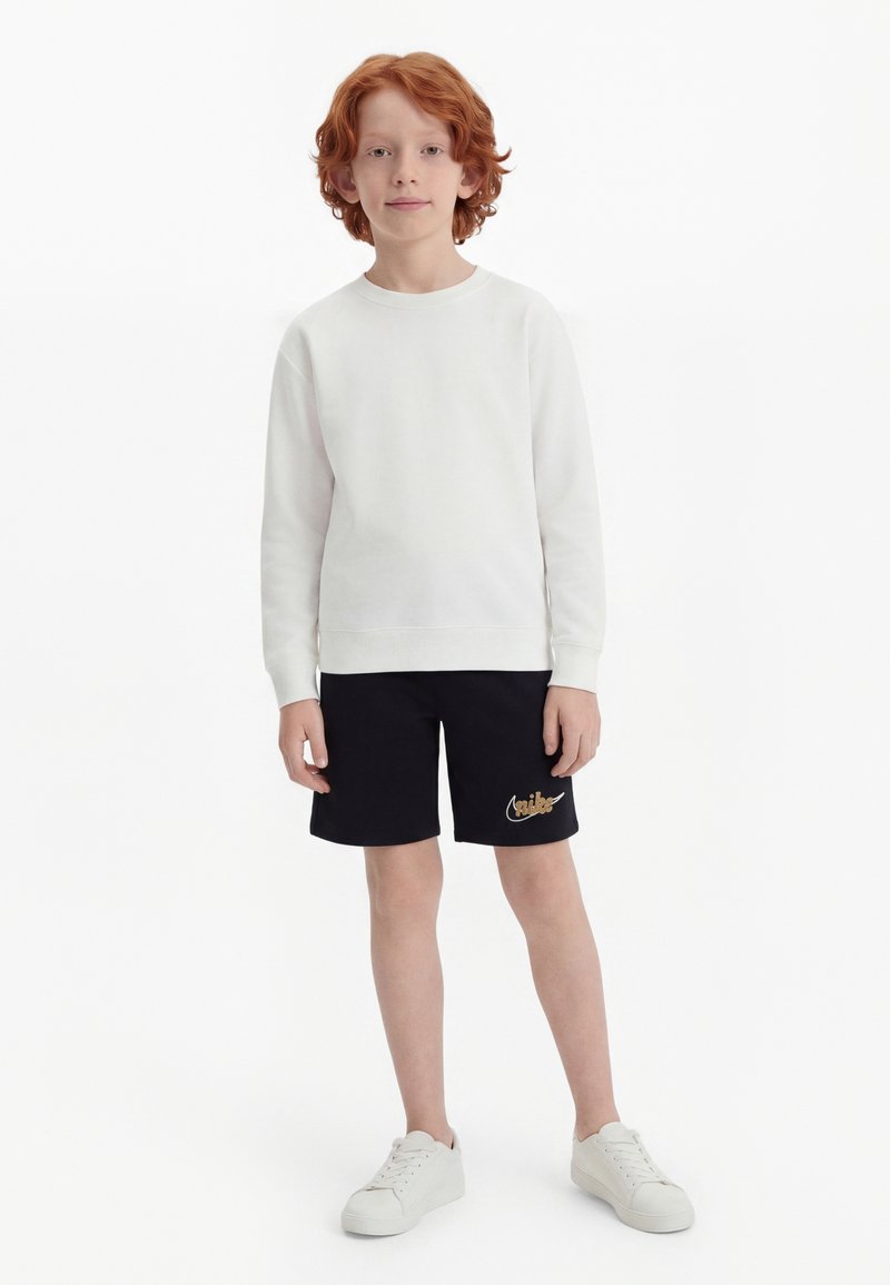 Παιδί με κόκκινα μαλλιά που φοράει λευκό φούτερ, μαύρη βερμούδα με χρυσό λογότυπο και λευκά αθλητικά παπούτσια, στέκεται μπροστά σε λευκό απλό φόντο.