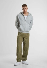 Grauer Zip-Hoodie mit Kapuze, vorderen Taschen, kombiniert mit olivgrünen, locker sitzenden Hosen und weißen Sneakers. Glatte Stofftextur.