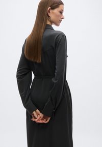 Μαύρο φόρεμα με μακριά μανίκια, εφαρμοστό στη μέση με ζώνη. Λείο ύφασμα, πλαϊνές σχισμές και γιακάς στο λαιμό. Η πίσω όψη αναδεικνύει το κομψό σχέδιο.