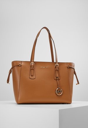 Bruine leren Michael Kors tote bag met dubbele handvatten, gouden logohanger en verstelbare zijriemen op een wit oppervlak.