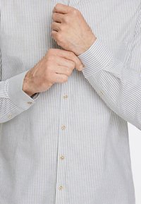 Camisa ligera a rayas con estrechas franjas gris y blancas, puños abotonados y botones de color marrón claro, que presenta un cuello clásico y una textura suave.