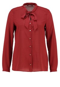 Blouse rouge à motifs avec manches longues, col rond et détail nœud papillon au niveau du col. Dotée d'une fermeture boutonnée et d'un tissu texturé.