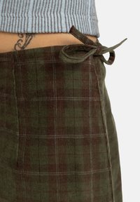Grön kordfloskjol med rutig mönstring och röda accenter. Midjan är knuten med en rosett. Synlig textur och sömnadsdetaljer.