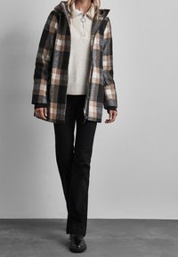 Rutig jacka i ullblandning i svart, beige och grå, med huva, dragkedja och sidofickor. Buren över en krämfärgad tröja och svarta byxor.