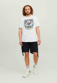 Man with curly hair wearing white graphic t-shirt, black shorts, white sneakers, standing against plain light background.