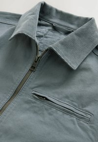 Veste bleu clair zippée avec un col texturé et une poche sur la poitrine. Fabriquée en coton avec des coutures visibles et une fermeture éclair en laiton.