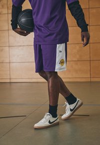 Canotta da basket in rete viola e pantaloni corti con logo dei Lakers, abbinati a calze nere a girocollo e sneakers bianche con swoosh nero.