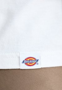 Bijela pamučna majica s vidljivom oznakom Dickies na rubu, postavljena na svijetlosmeđoj pozadini. Glatka tekstura.
