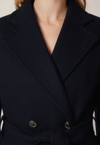 Granatowy płaszcz dwurzędowy z wyraźnymi klapami, paskiem w talii i srebrnymi guzikami; wykonany z teksturowanej tkaniny o gładkim wykończeniu.