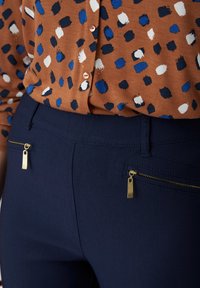 Pantaloni navy con dettagli in zip dorati, abbinati a una blusa marrone con un motivo astratto a macchie colorate in blu, bianco e nero.