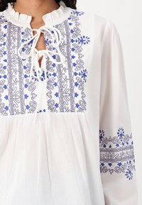 Blouse blanche avec broderies bleu et marine présentant des motifs floraux et en spirale. Encolure froncée et détail à nouer à l'avant. Coupe ample.