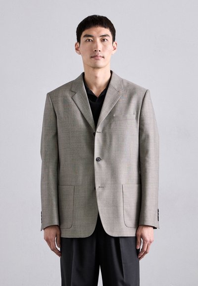 Blazer texturizado de color gris claro, con solapas de muesca, dos bolsillos frontales y cierre de tres botones. Llevado sobre una camisa negra con un corte ajustado.