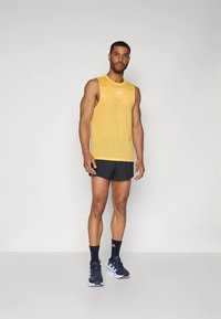 Camisola de alças amarela em tecido rede com logótipo, combinada com calções pretos e ténis desportivos azuis escuros, em pé sobre um fundo neutro.