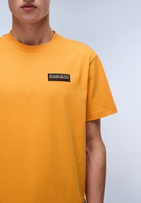 Žluté krátké tričko s krátkým rukávem vyrobené z bavlny, s obdélníkovou černou logotypovou nášivkou na levé části hrudi. Hladká textura, design s kulatým výstřihem.