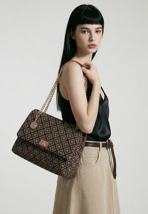 Jeune femme aux cheveux noirs et raides, portant un top en satin noir et une jupe beige, tenant un grand sac à main marron à motifs avec des anses en chaîne dorée.