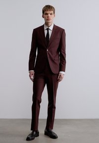 Κοκκινωπό κοστούμι σε στενή γραμμή, με κούμπωμα ενός κουμπιού, πέτο με εγκοπή και μαύρη γραβάτα, συνδυασμένο με μαύρα παπούτσια σε απλό φόντο.