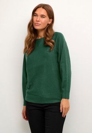 Grüner Strickpullover mit lockerer Passform, Rundhalsausschnitt und gerippten Bündchen sowie Saum. Weiche Textur und leicht übergroßes Design. Getragen mit schwarzen Hosen.