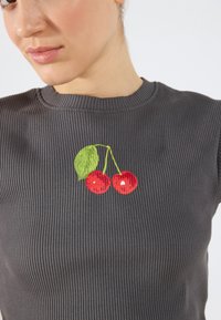 Mørk grå ribbet topp med rød kirsebærbroderi og grønne blader i sentrum. Stoffet har en teksturert overflate.