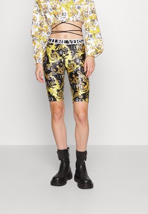 Femme portant un short cycliste noir et or à motifs, un top court assorti à manches longues, des bottes noires à lacets sur un fond clair.