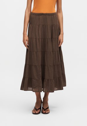 Mallilla on päällään ruskea kerroksittainen midihame, oranssi hihaton paita ja mustat korolliset sandaalit, ja hän seisoo yksiväristä valkoista taustaa vasten.