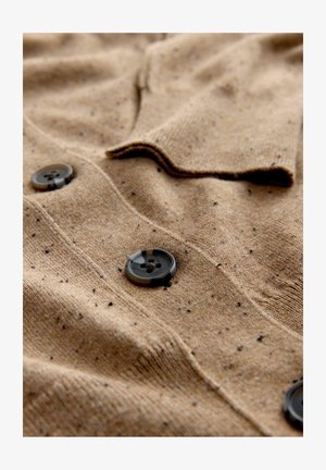 Beige kudumüts, millel on tekstuuriga kangas, millel on täpikestega detailid ja mustad nööbid. Sel on lähedane krae ja eesmine nööbiga sulgemine.