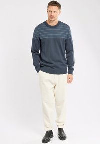 Pull en maille bleu marine avec des rayures texturées horizontales, col rond, poignets et ourlet côtelés. Associé à un pantalon beige et des bottes noires.