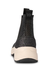 Zapatillas negras sin cordones con un respaldo texturizado, que presentan un material brillante y una suela de color crema para crear contraste. Dos tiras en la parte trasera para facilitar su colocación.