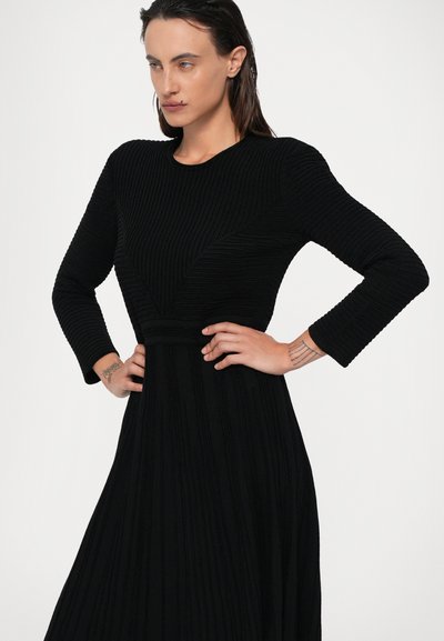 Vestido preto de manga longa com nervuras, com um corpo justo e saia rodada. Possui um decote redondo e um padrão texturizado por todo o vestido.