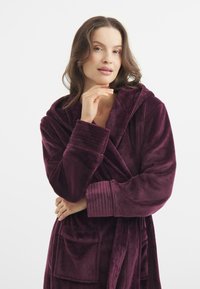 Accappatoio di velluto, di un profondo colore viola, con cappuccio, polsini a coste e tasca frontale; la sua texture morbida e il design accogliente sono caratteristiche distintive.
