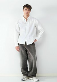 Weiß gestreiftes Hemd mit Knöpfen, graue weite Jeans und hellgraue Turnschuhe. Das Model steht vor einem neutralen Hintergrund.