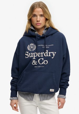 Lyshåret kvinde iført navy blå Superdry hættetrøje over en stribet skjorte og hvide bukser, stående mod hvid baggrund.