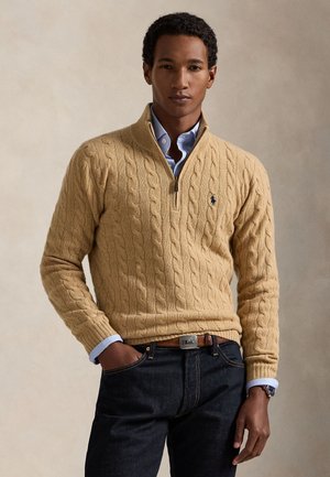 Homme portant un pull beige en maille torsadée à fermeture éclair quart, par-dessus une chemise bleue avec un jean foncé, debout avec une main dans la poche devant un fond uni.