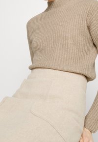 Pull beige en tricot avec un col montant et une texture côtelée, associé à une jupe beige clair avec des poches et un tissu doux.