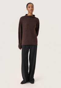 Brauner Strickpullover mit hohem Kragen, langen Ärmeln und lockerer Passform, kombiniert mit schwarzen weiten Hosen und schwarzen Schuhen.