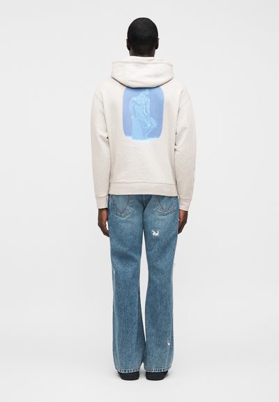 Bež hudica s grafikom na leđima u obliku plave siluete statue, uparena sa širokim trapericama plave boje s oštećenjima. Minimalistički dizajn, teksturirani materijal.